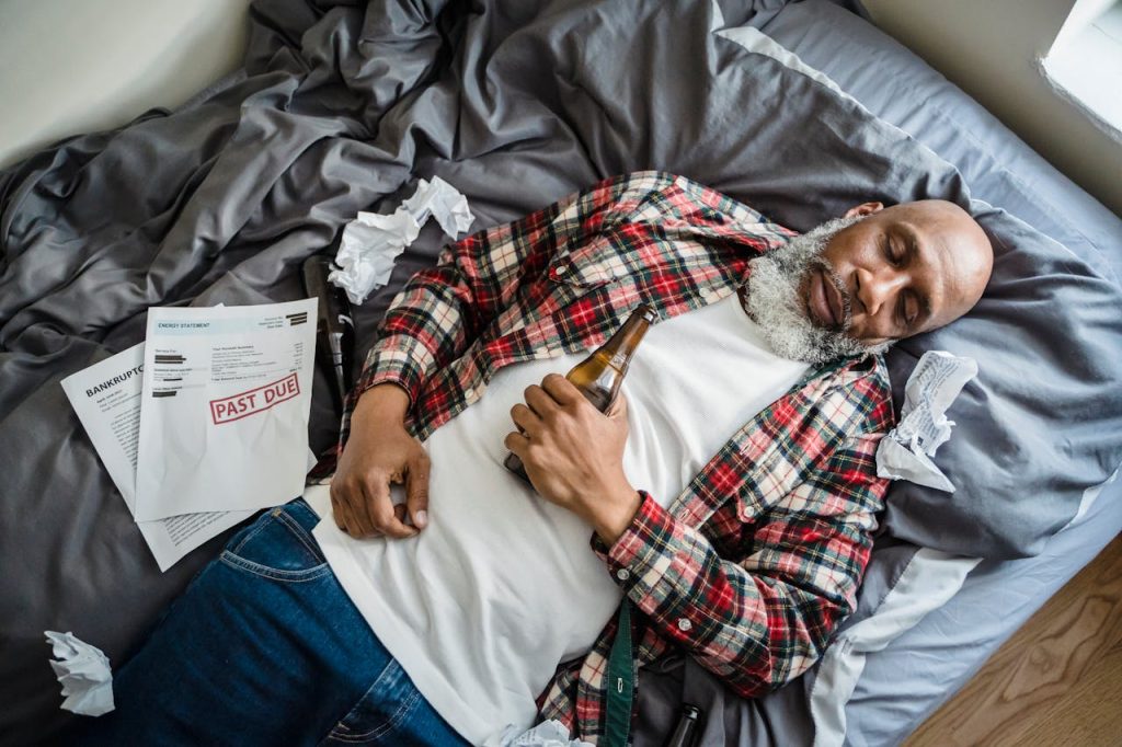 A senior man asleep on a bed, surrounded by past due bills, holding a beer bottle.