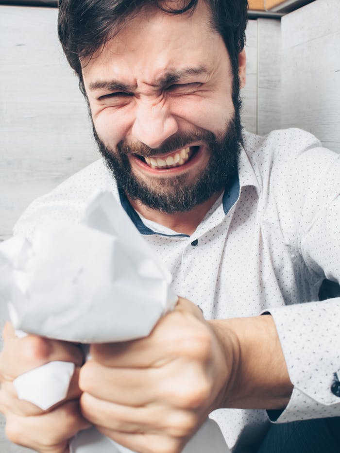 A stressed bearded man crumples paper in frustration indoors, showing intense emotion.